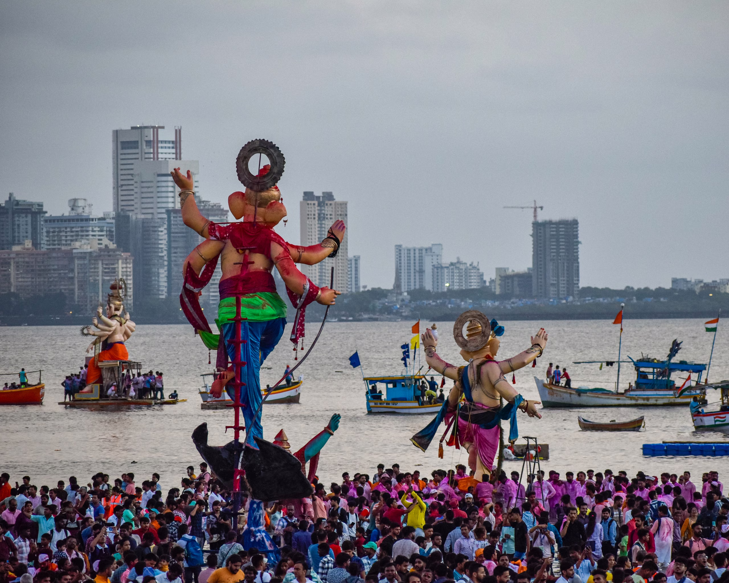 Large painted Ganesha idols carried through a crowd toward the sea, fishing boats offshore, high-rise city skyline behind under grey sky