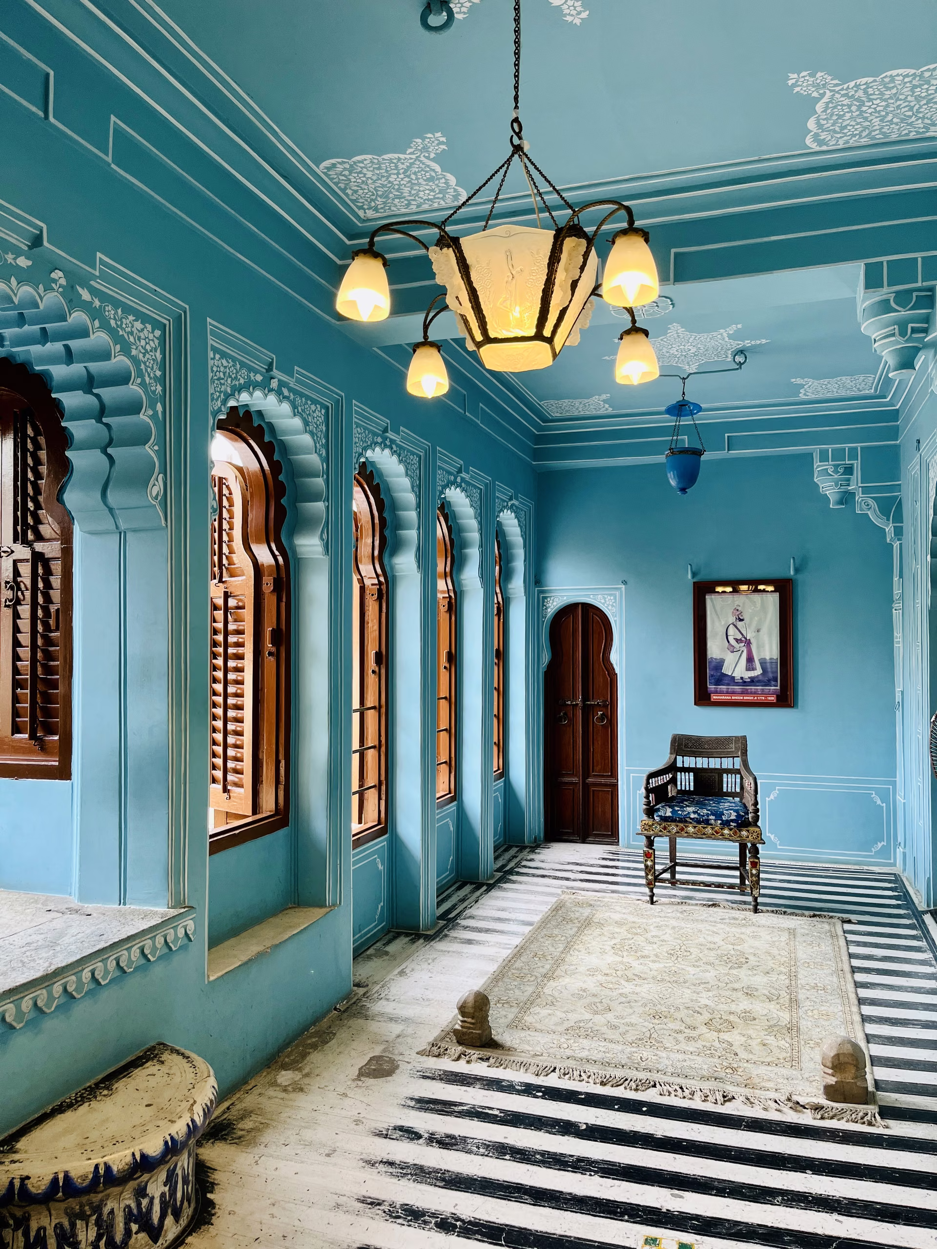 Turquoise painted palace room with cusped arch windows, wooden shutters, glass chandelier, carved chair, framed portrait, striped floor and rug.