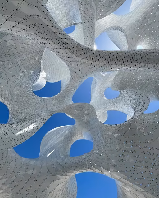 Upward view of a perforated white organic-shaped canopy structure with intricate dot patterns, revealing circular and oval openings to a clear blue sky.