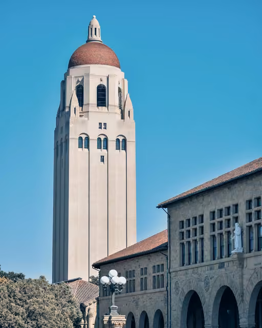 Tall cream sandstone bell tower with a red-tiled dome, next to an arched building with a statue, under a clear blue sky.