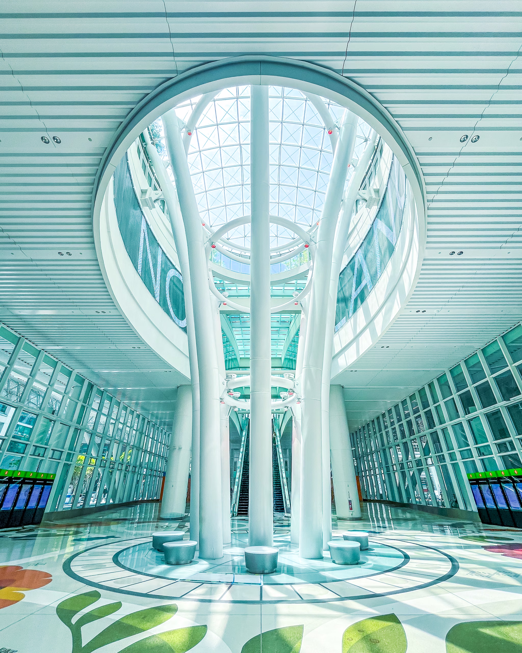 Symmetrical station atrium with white branching columns rising to a circular glass skylight, escalators behind, floral floor graphics