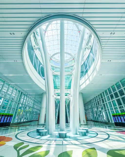 Symmetrical station atrium with white branching columns rising to a circular glass skylight, escalators behind, floral floor graphics