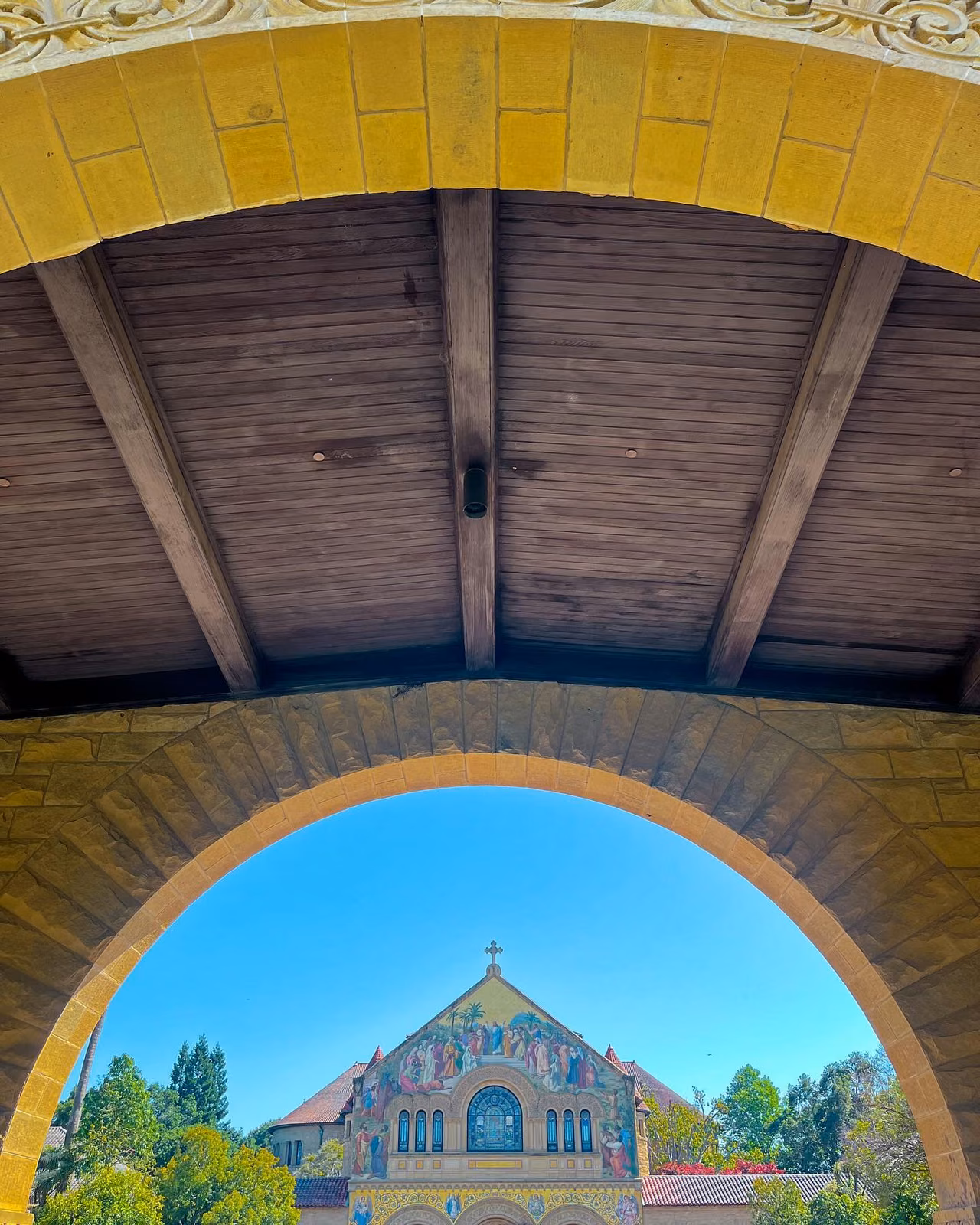 View through a stone archway and under a wooden beamed ceiling toward a mosaic-decorated church facade with a cross, framed by trees.