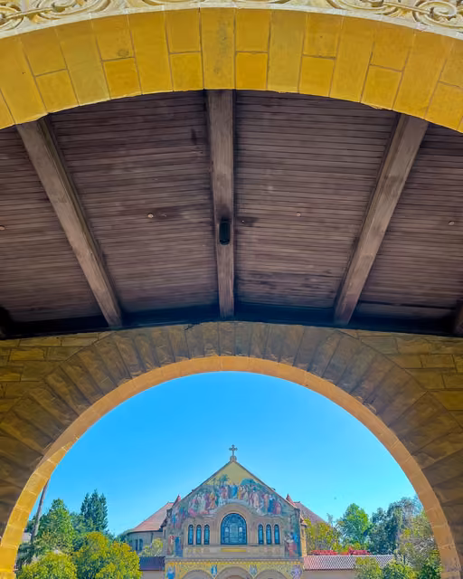 View through a stone archway and under a wooden beamed ceiling toward a mosaic-decorated church facade with a cross, framed by trees.