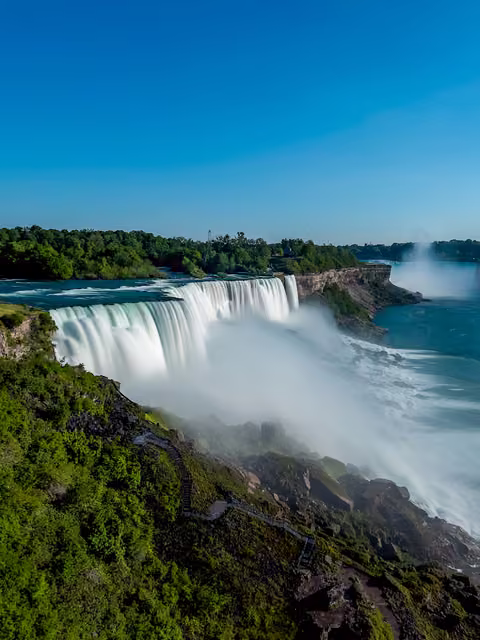 Wide waterfall with long-exposure smooth white water plunging over a curved rock ledge, mist rising, green cliffs and blue sky