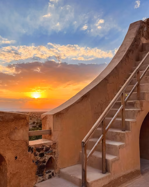 Curved mudbrick staircase with wooden handrails leads up a sandy-colored fortress wall, silhouetted against an orange sunset sky over a desert landscape.