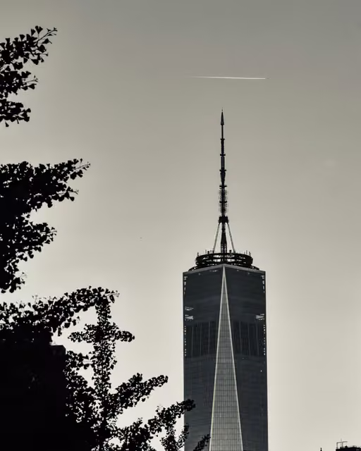 Upper floors and antenna spire of a tall glass skyscraper against a pale sky, with silhouetted ginkgo tree branches at left and a jet contrail overhead.