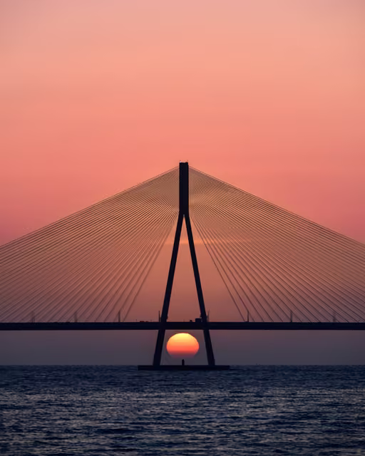 Silhouetted cable-stayed bridge pylon over water with the setting sun framed between its two legs beneath the deck.