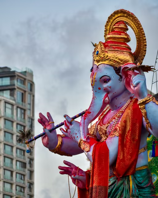 Large blue Ganesha idol playing a flute, wearing gold crown, red silk drape and pearl necklaces, against overcast sky beside a high-rise apartment building