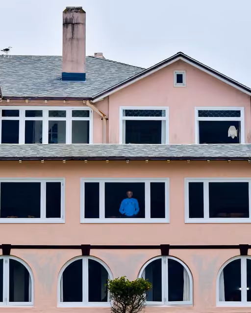 Pink multi-story building facade with a chimney, gabled roof, and a man in a blue sweater standing at a middle-floor window.