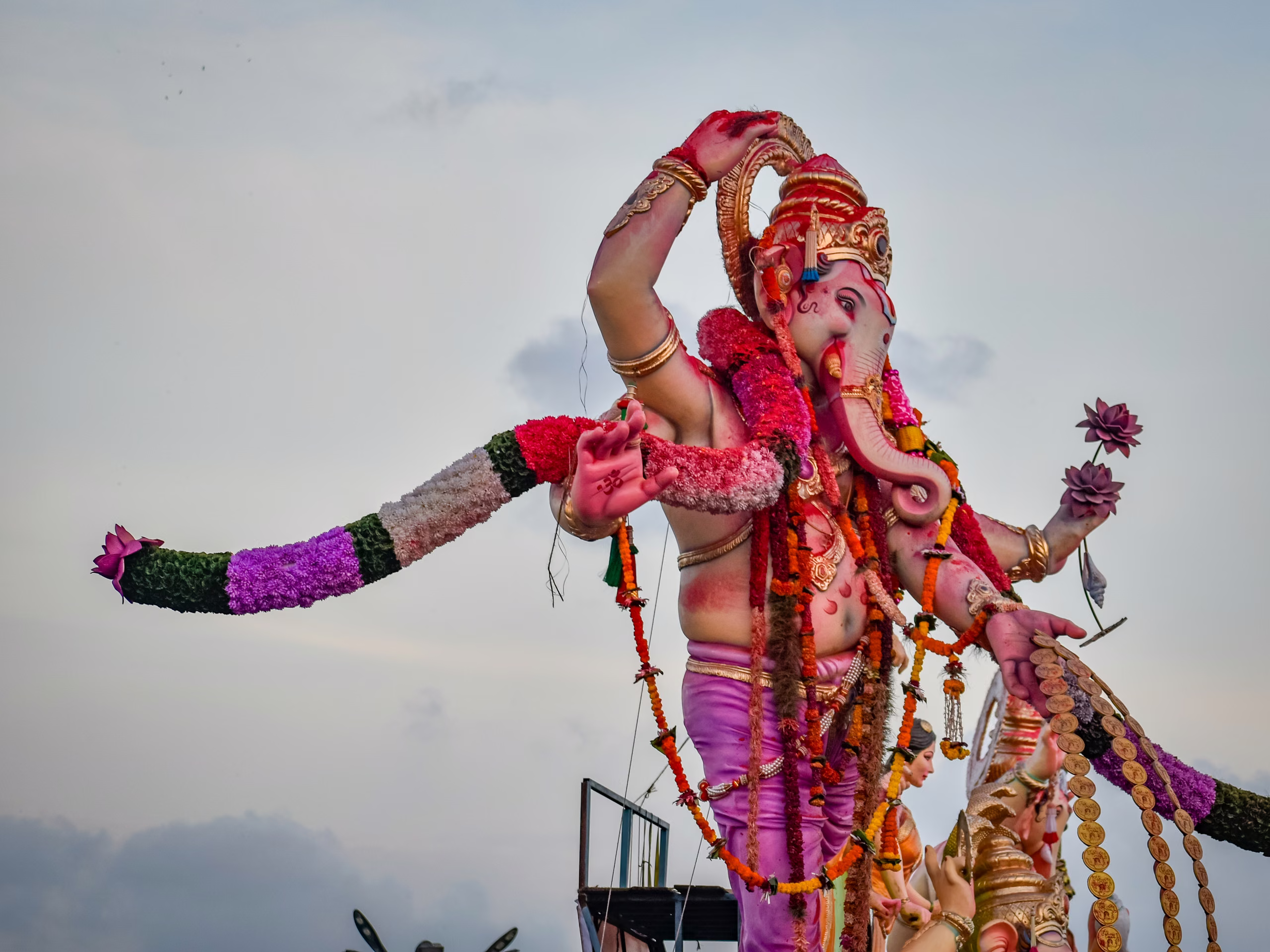 Large pink-skinned Ganesha idol with gold crown, marigold garlands and flower-wrapped arms, raised against pale overcast sky