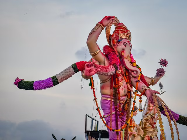Large pink-skinned Ganesha idol with gold crown, marigold garlands and flower-wrapped arms, raised against pale overcast sky