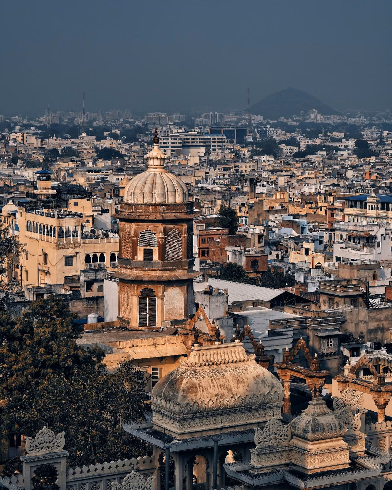 Aerial view of a domed palace tower with ornate carved cupolas in the foreground, overlooking a dense cityscape of rooftops stretching to a hazy hill.