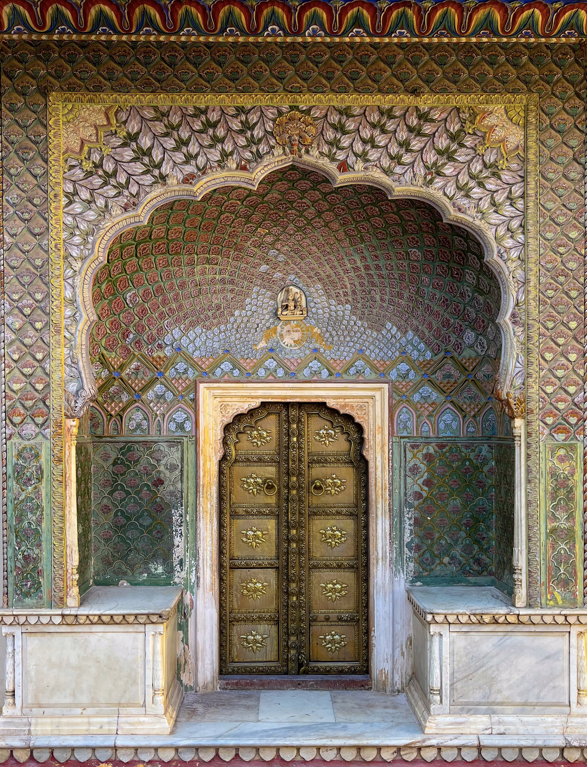 Ornate arched palace doorway with brass studded double doors, rose-patterned painted plasterwork, marble benches either side and stone floor