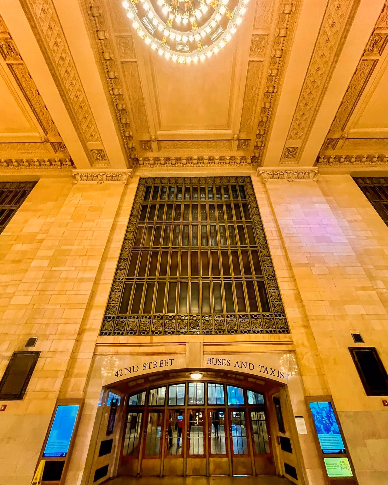 Ornate marble entrance hall with tall arched window grille, carved ceiling, chandelier, and doors labeled '42nd Street Buses and Taxis'.