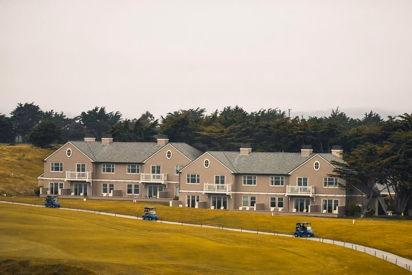 A large tan shingled clubhouse with white balconies sits along a golf course, with three blue golf carts on the cart path and tall trees behind.