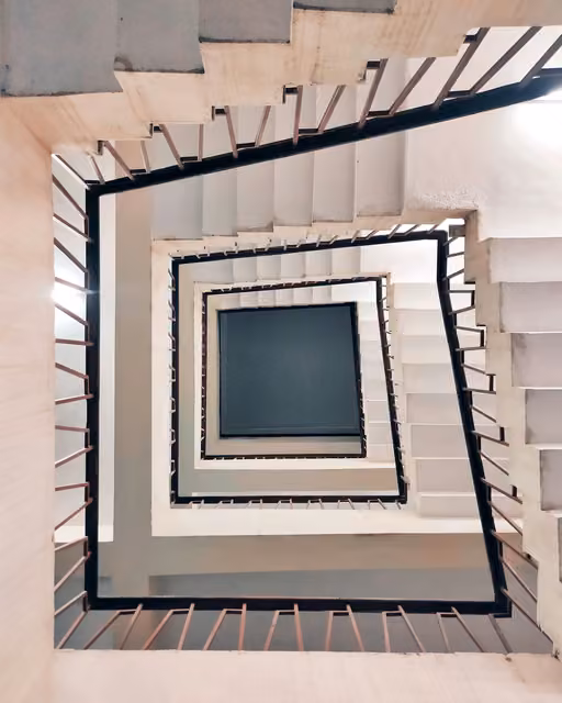 Looking straight down into a square spiral staircase with white steps and black-outlined railings, forming concentric squares toward a gray floor below.