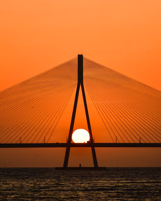 Silhouetted cable-stayed bridge pylon over water, the sun setting directly between its two angled legs against an orange sky