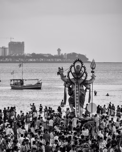 Black-and-white view of a large Ganesh idol carried into the sea, surrounded by a dense crowd, with a boat and city skyline behind.