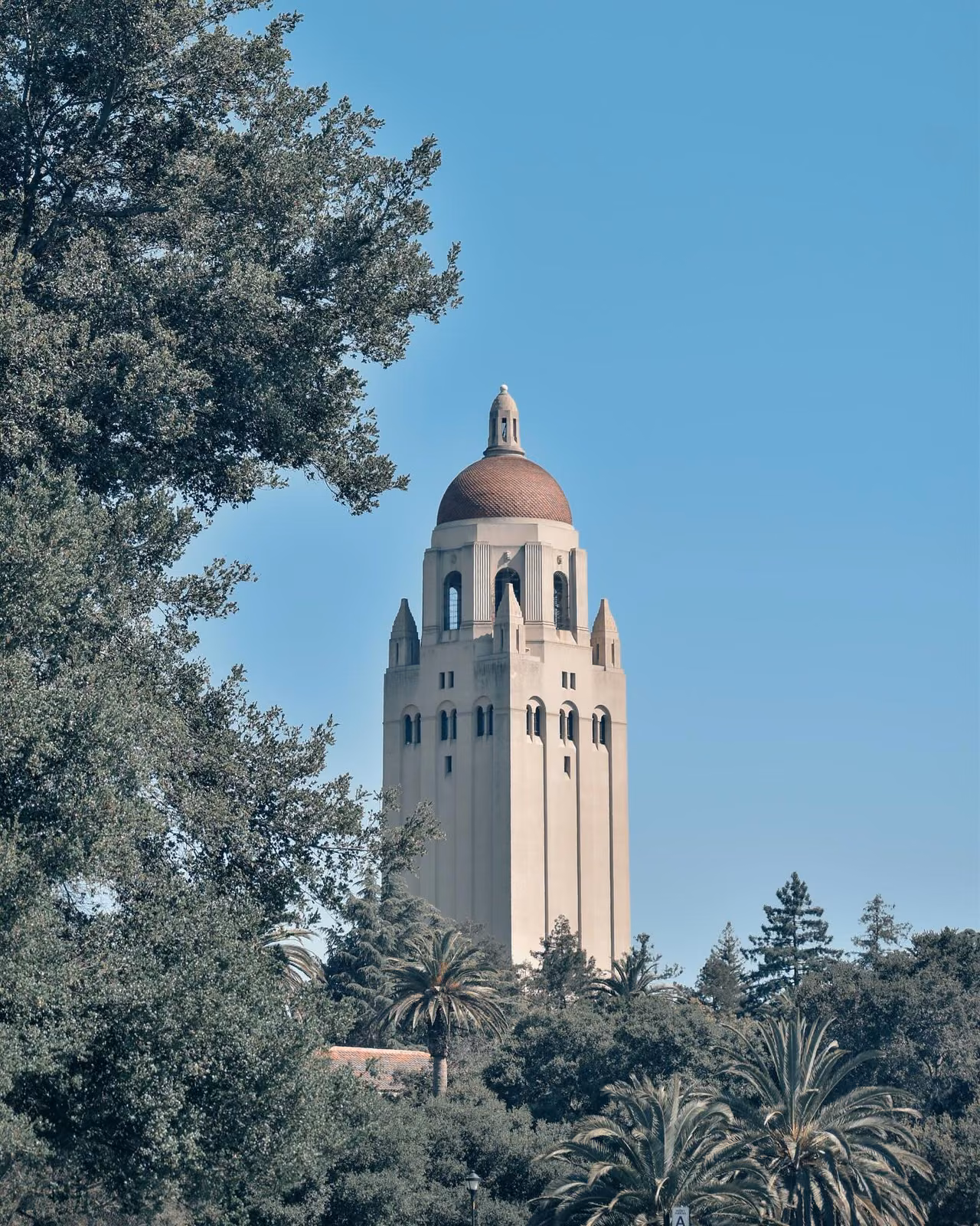 A tall cream-colored tower with a domed reddish-brown roof rises above surrounding green trees under a clear blue sky.