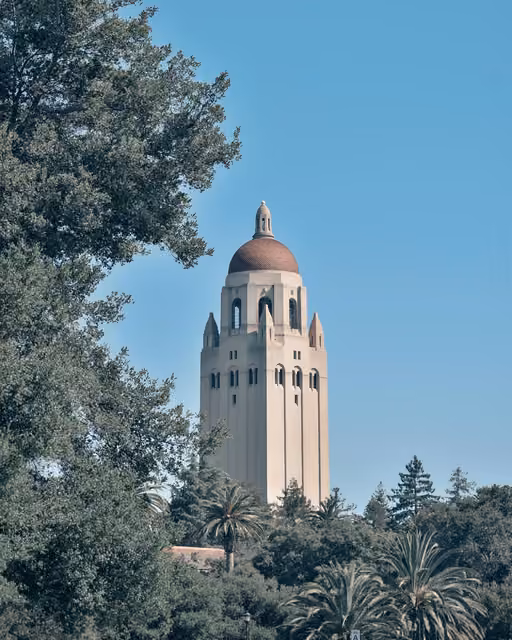 A tall cream-colored tower with a domed reddish-brown roof rises above surrounding green trees under a clear blue sky.