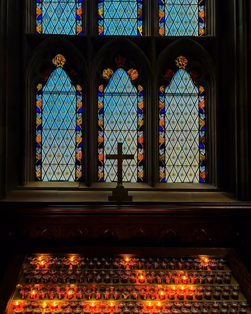 Three Gothic stained-glass windows above a wooden altar rail, with a dark cross in front and rows of lit red votive candles below.