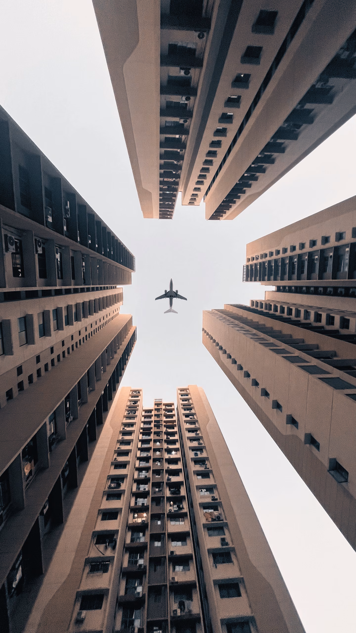 Upward view from a courtyard between four tan high-rise apartment blocks, with an airliner crossing the pale sky above