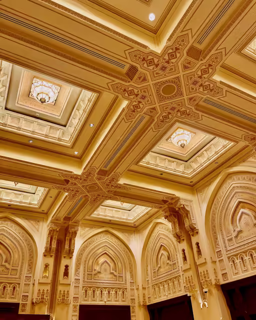 Ornate coffered ceiling in cream and gold with geometric Islamic patterns, recessed skylight panels, hanging lanterns, and pointed arch wall niches below
