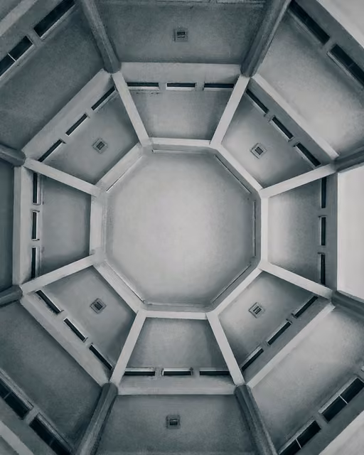 Symmetrical view looking straight up into an octagonal atrium ceiling with radiating white beams and small skylight vents, in monochrome.