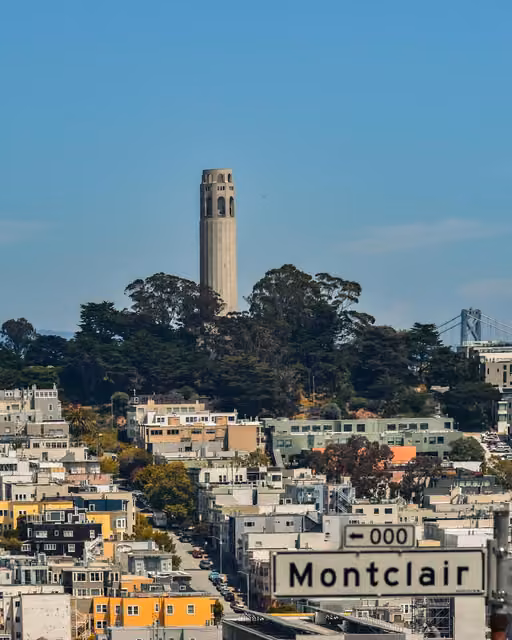 Coit Tower rises above tree-covered hill overlooking San Francisco rooftops, with a street sign reading Montclair in foreground and a bridge visible at right.