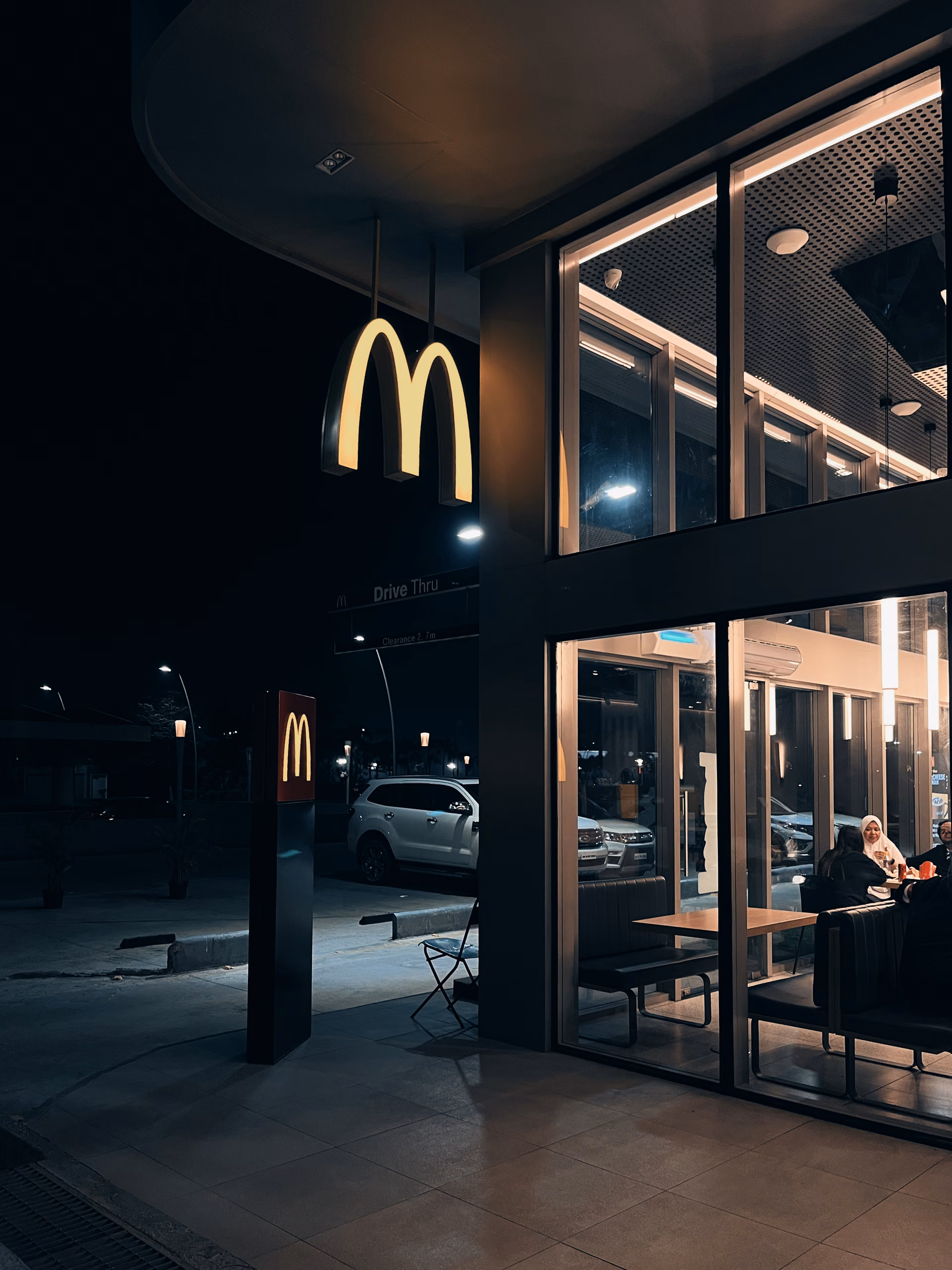 Illuminated McDonald's golden arches sign at night outside a glass-fronted restaurant, two women seated inside at a table.