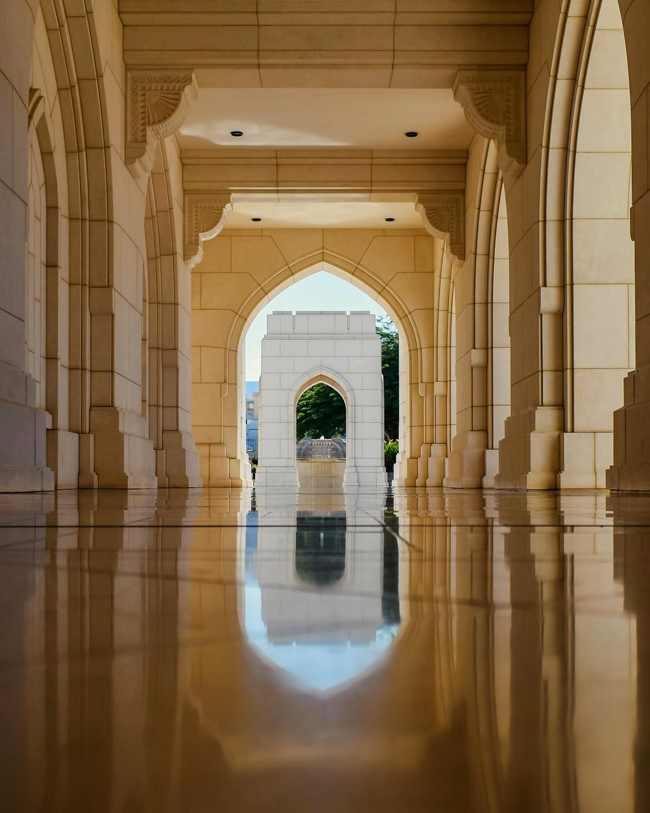 A polished marble corridor with pointed Islamic-style arches leads to an outdoor archway framing a fountain and trees, with reflections visible on the wet floor.