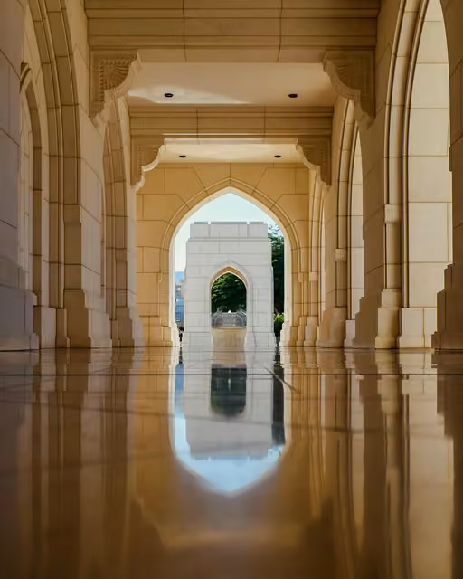 A polished marble corridor with pointed Islamic-style arches leads to an outdoor archway framing a fountain and trees, with reflections visible on the wet floor.