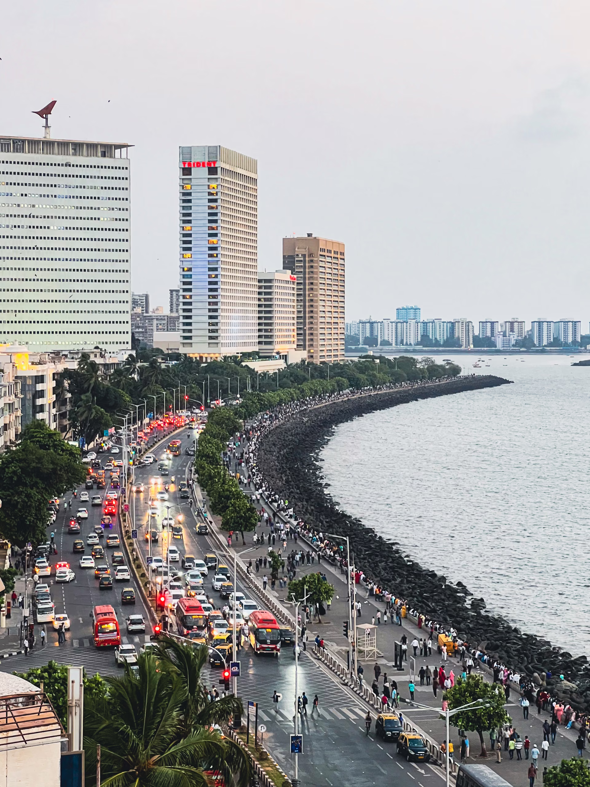 Elevated view of a curving seafront boulevard with heavy traffic, crowds on the promenade, tetrapod breakwater and high-rise buildings at dusk