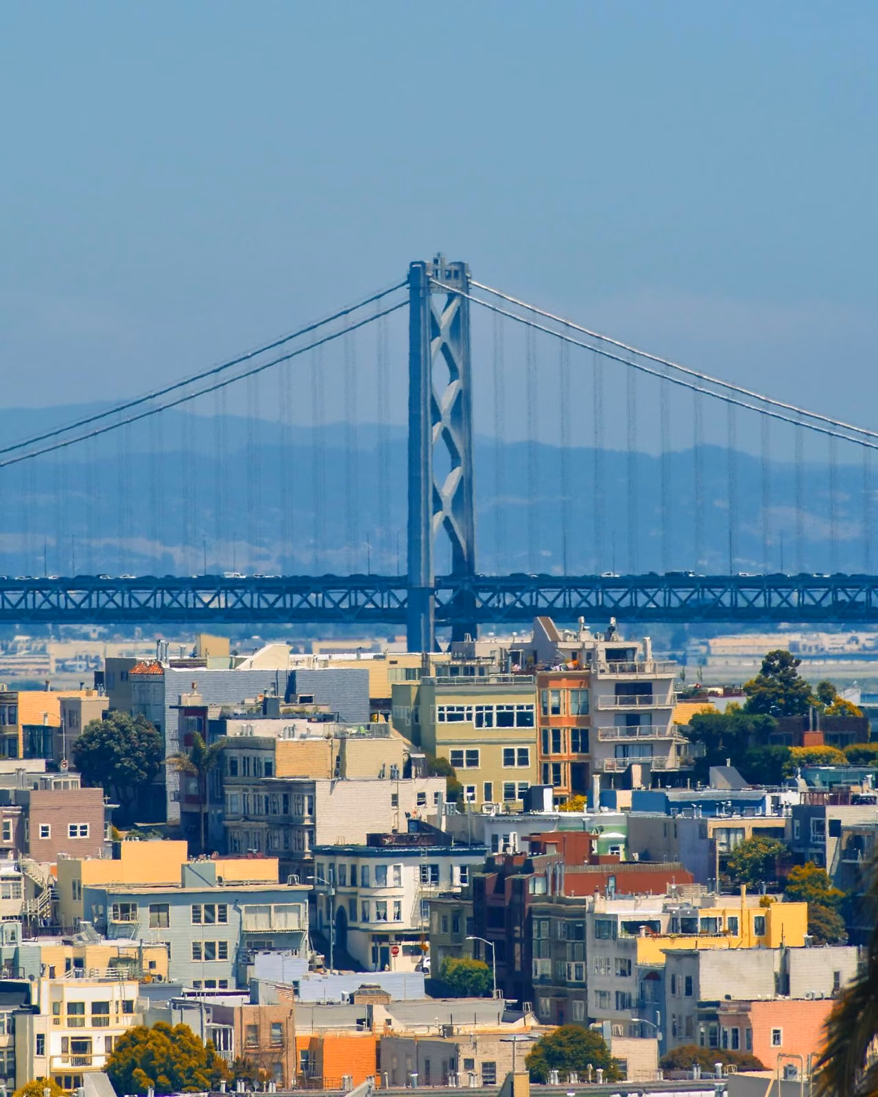 View of the San Francisco–Oakland Bay Bridge suspension tower rising above colorful residential rooftops, with hazy hills in the background under a clear sky.