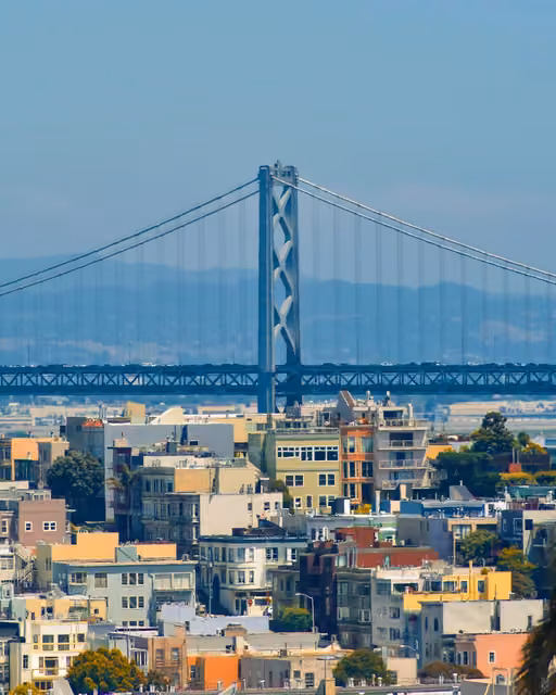 View of the San Francisco–Oakland Bay Bridge suspension tower rising above colorful residential rooftops, with hazy hills in the background under a clear sky.