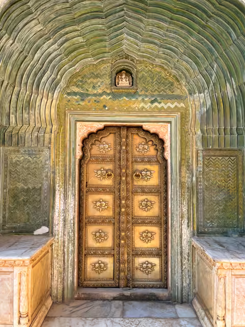 Ornate gold-painted double door with floral bosses and ring handles, set in a green scalloped arch with marble benches flanking it